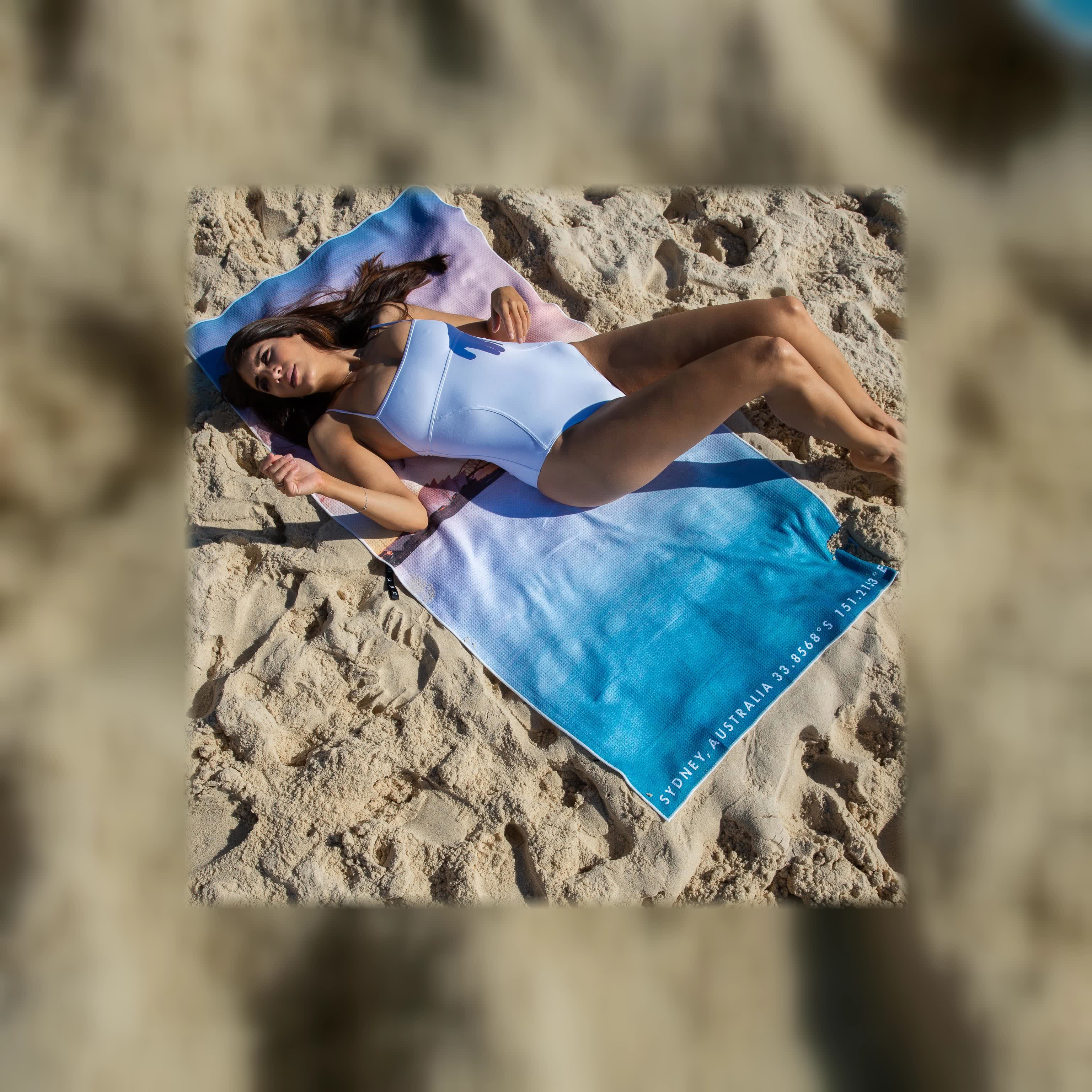 A beach towel on the sand shot from above, its printed GPS coordinates running along the edge: Sydney, Australia