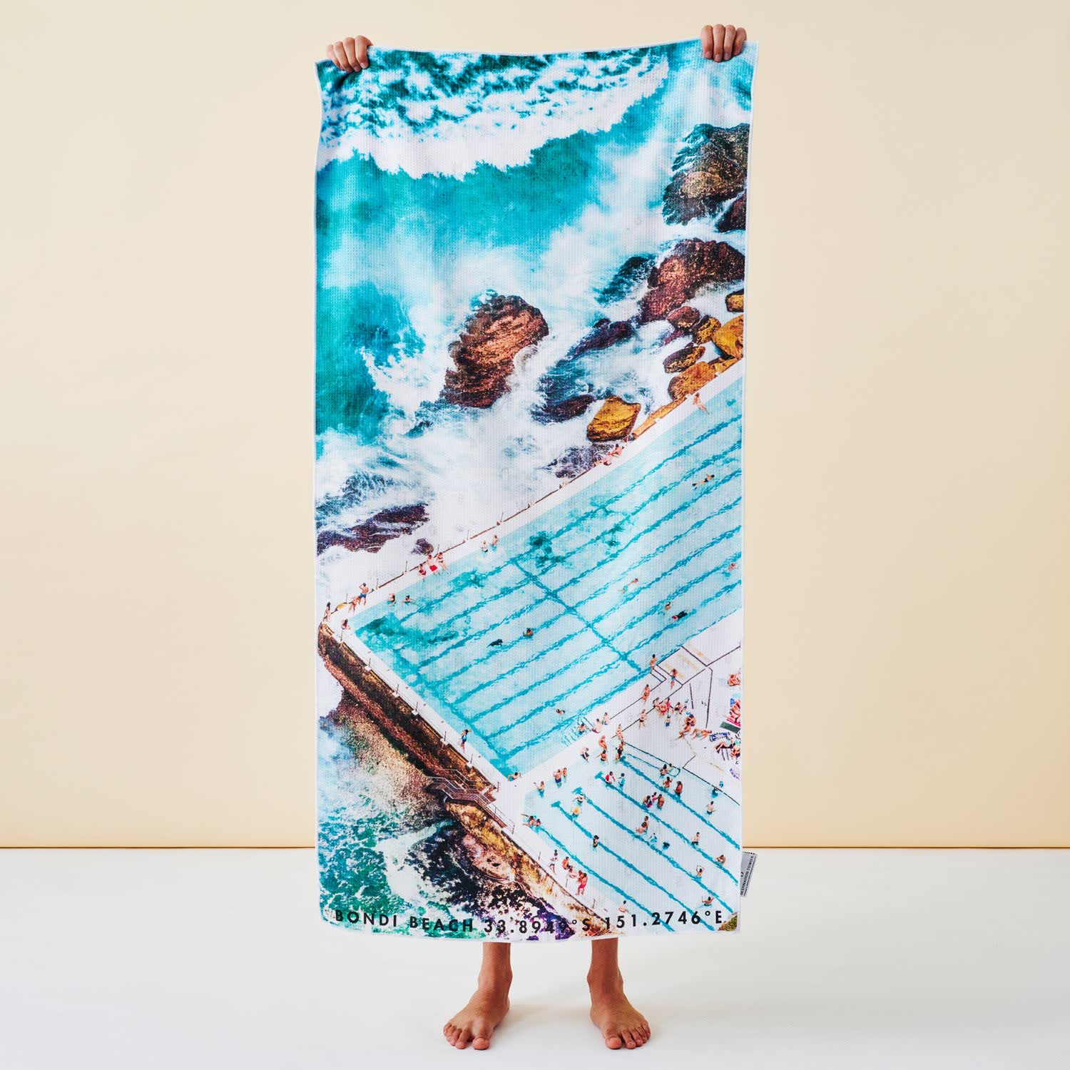 The Icebergs Summer beach towel held up, printed edge to edge with an aerial photograph of Bondi Icebergs